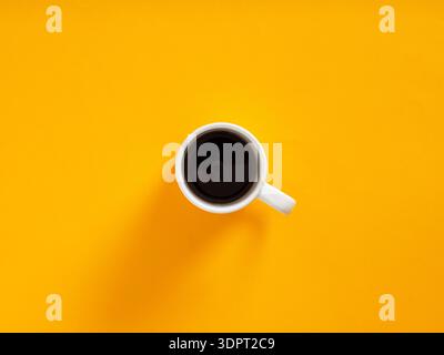 Tazza da caffè bianca riempita di bevanda nera mattutina, che crea un contrasto minimo su una superficie gialla vibrante Foto Stock