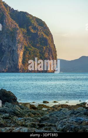 Una ripida isola rocciosa di pietra calcarea sorge da calme acque marine tropicali, vista da una costa rocciosa con pietre lisce in primo piano. Foto Stock