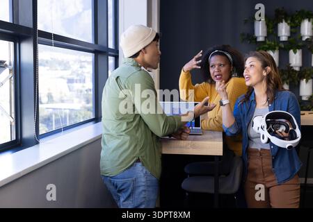 Diversi colleghi seduti intorno a un tavolo in legno alto in ufficio che discutono di visore VR e laptop Foto Stock