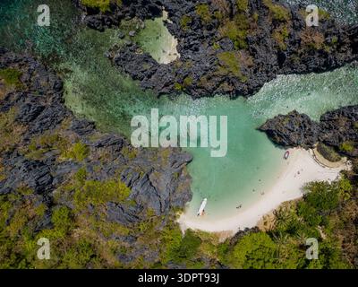 Vista aerea dall'alto verso il basso di una piccola spiaggia di sabbia circondata da formazioni rocciose di pietra calcarea scura e acque turchesi poco profonde che formano una stretta laguna. Foto Stock