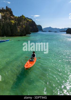 Una persona pagaiando in kayak attraverso le acque verdi cristalline in una laguna calcarea riparata, con scogliere ripide e piccole barche visibili in lontananza. Foto Stock