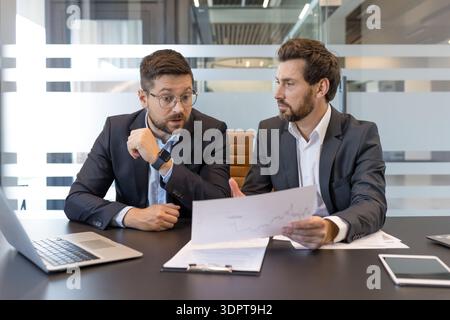 Due uomini d'affari in tuta discutono documenti finanziari in un moderno tavolo da ufficio, collaborano e analizzano i dati di mercato durante la pianificazione della strategia aziendale durante una riunione Foto Stock