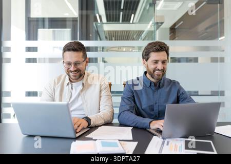 Due colleghi maschi sorridenti che collaborano a una scrivania moderna e luminosa, lavorano su computer portatili e documenti, pianificano un progetto aziendale con attenzione, lavoro di squadra ed energia positiva Foto Stock