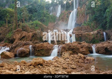 Un'ampia cascata a più livelli scorre su formazioni rocciose marroni stratificate in un fiume poco profondo, circondato da una fitta foresta tropicale. Foto Stock
