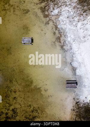 Una vista verticale dall'alto verso il basso mostra due tavoli da picnic in legno parzialmente sommersi dall'acqua poco profonda del lago accanto a una costa di sabbia pallida. Foto Stock