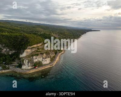 Una strada costiera curva lungo ripide scogliere marine accanto alle calme acque dell'oceano all'alba, con colline boscose che si estendono lungo la costa. Foto Stock