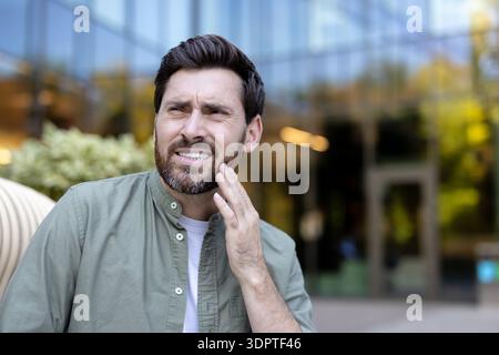 Uomo che soffre all'aperto, che soffre la guancia dal mal di denti, mostra disagio dentale e urgenza per l'assistenza dentale, uomo barbuto di mezza età che guarda lontano alla luce del giorno Foto Stock