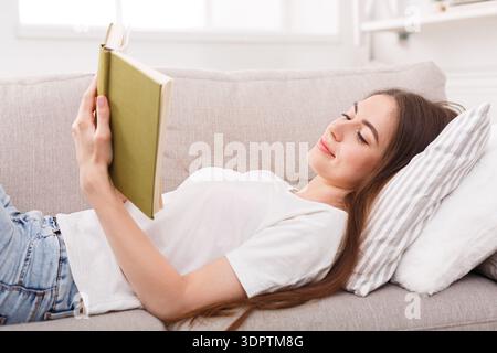 Una giovane donna ama leggere un libro su un divano accogliente in una stanza luminosa durante il pomeriggio Foto Stock