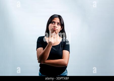 Seria donna del sud-est asiatico in t-shirt nera che guarda con attenzione con il dito sul mento. Studio isolato girato su sfondo bianco, idea concettuale. Foto Stock