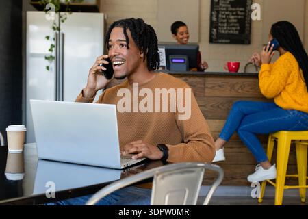 Uomo afroamericano che scrive sul computer portatile, tiene lo smartphone al tavolo del bar con una tazza di carta Foto Stock