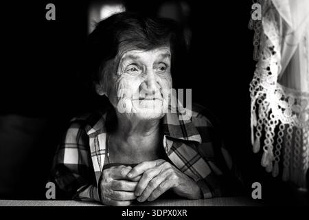Ritratto in bianco e nero di una donna anziana seduta accanto a una finestra. La luce naturale soffusa mette in risalto il suo viso e le sue mani espressive, creando un'atmosfera intima Foto Stock