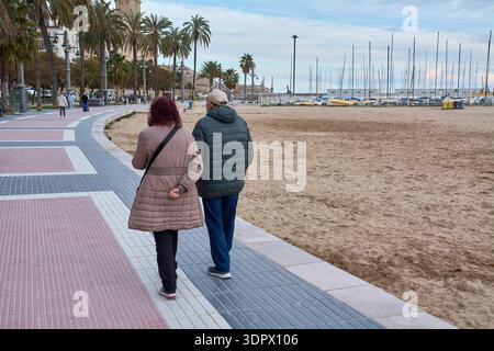 Sitges. Spagna - 15 febbraio 2026: Coppia senior che cammina lungo il lungomare vicino alla chiesa e al porticciolo in una tranquilla giornata invernale in questo coa mediterraneo Foto Stock