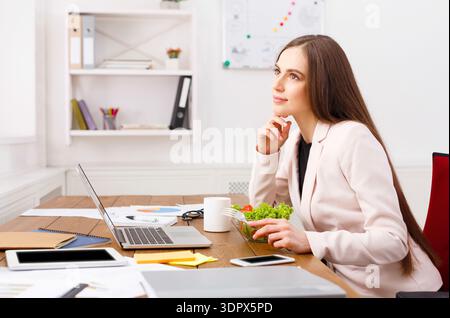 Donna che pensa mentre è seduta alla scrivania con insalata e laptop in ufficio Foto Stock