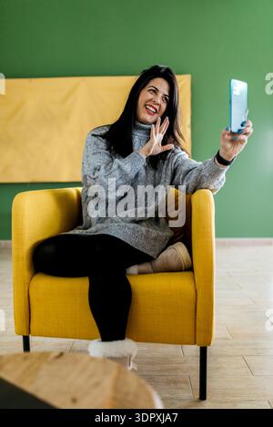 Una donna seduta su una comoda poltrona gialla scatta un allegro selfie con il suo smartphone. Indossando un maglione grigio, sorride calorosamente, godendosi un mo rilassato Foto Stock