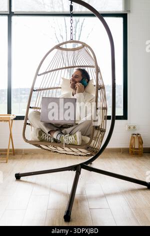 Uomo sorridente che lavora da casa seduto a gambe incrociate in una comoda sedia sospesa. Parla al telefono e gode di un momento di produttività in Foto Stock