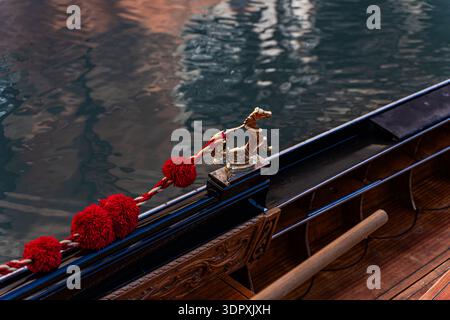 Primo piano di una gondola veneziana che mostra intricate sculture in oro e dettagli in corda rossa. Il riflesso degli edifici nell'acqua mette in risalto la Venezia Foto Stock