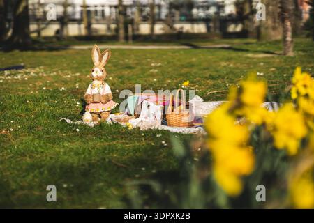 Un incantevole picnic pasquale in un parco soleggiato presenta una statuina di coniglietto pasquale vestito e un cestino di vimini su una lussureggiante erba verde con narcisi nel Foto Stock