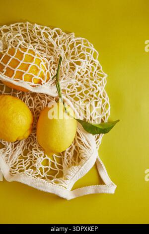 Limoni gialli luminosi disposti in un sacchetto di rete riutilizzabile su uno sfondo giallo brillante, che mette in risalto freschezza e sostenibilità nelle scelte alimentari attraverso vi Foto Stock
