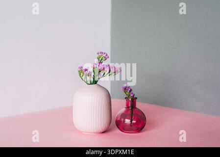 Due piccoli vasi con delicati fiori viola si trovano elegantemente su una superficie rosa su uno sfondo grigio e bianco a due tonalità, creando un design minimalista Foto Stock