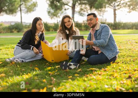 Un gruppo di studenti diversi seduti sull'erba, godendo di una sessione di studio all'aperto. Sono impegnati con un tablet e materiali, circondati dalla natura Foto Stock
