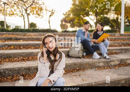 Gli studenti si godono una soleggiata giornata autunnale all'aperto. Una giovane donna a fuoco sorride mentre parla al telefono, con due compagni di classe che chiacchierano e studiavano Foto Stock