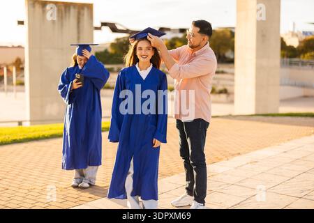 Un gioioso momento di laurea mentre gli amici festeggiano insieme. Una giovane donna con un abito blu ha il berretto regolato da un amico, mentre un'altra cattura il mo Foto Stock