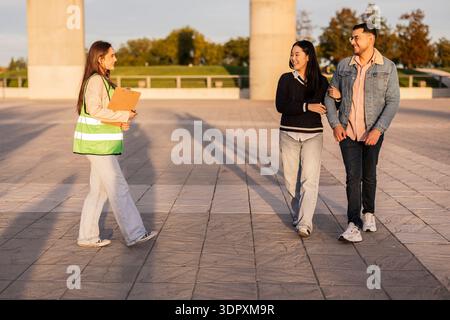 Una giovane donna con gli appunti sorride mentre si avvicina a due studenti che camminano braccio in braccio fuori in una giornata di sole. Si impegnano calorosamente in un ambiente spazioso Foto Stock