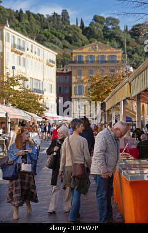 Gli amanti dello shopping curiosano tra le bancarelle di un mercato all'aperto a Nizza, nel sud della Francia, con edifici pastello e verdi colline dietro. Foto Stock