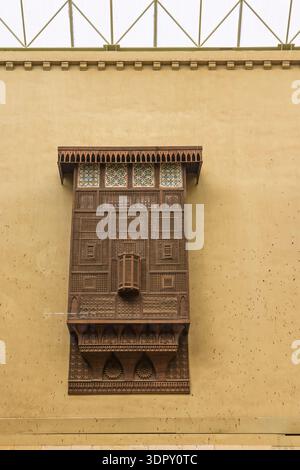 Finestra ornata in legno mashrabiya con intricato reticolo geometrico e vetrate colorate adagiate su una calda facciata gialla, che mostra la tradizionale islamia egiziana Foto Stock