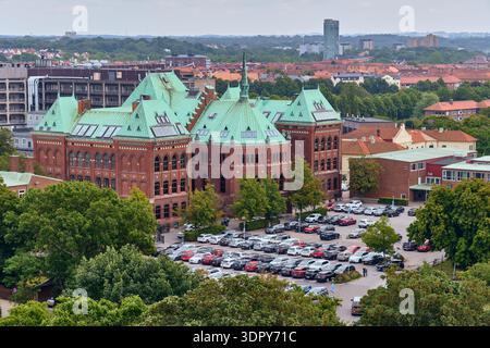 Helsingborg, Svezia - 12 agosto 2025: Veduta dei tetti verdi di rame di un grande edificio in mattoni rossi tra alberi lussureggianti e un parcheggio completo, nuovamente allestito Foto Stock