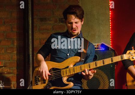 Presentazione Babsea CD musicista maschile che suona il basso elettrico sul palco in un piccolo locale, esibizione espressiva con sfondo a parete in mattoni, a. Foto Stock