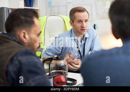 Uomo caucasico di mezza età seduto alla scrivania per discutere i dettagli del progetto con due colleghi maschi in ufficio, gesti con le mani, giubbotto di sicurezza e progetti visibili in background Foto Stock