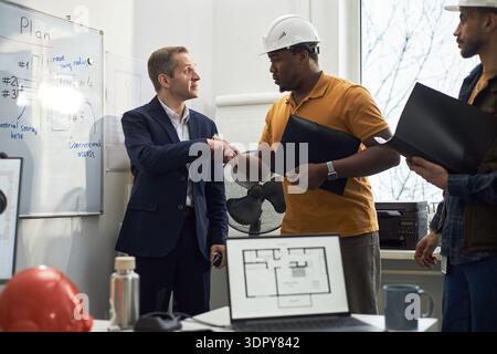 Uomo caucasico di mezza età in tuta da lavoro che stringe la mano a un uomo nero di mezza età che indossa un elmetto in ufficio, un team diversificato nelle vicinanze che tiene documenti, piani architettonici visibili Foto Stock