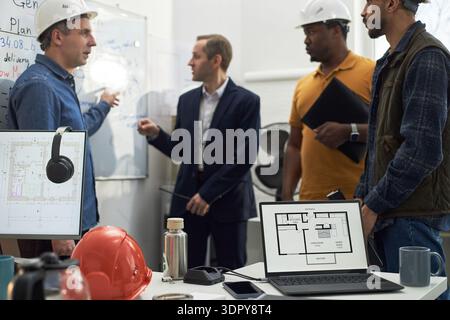 Gruppo di uomini di mezza età di diverse etnie che discutono i piani di costruzione in ufficio, un uomo caucasico che indica la lavagna mentre altri ascoltano, laptop che mostra la planimetria dell'edificio Foto Stock
