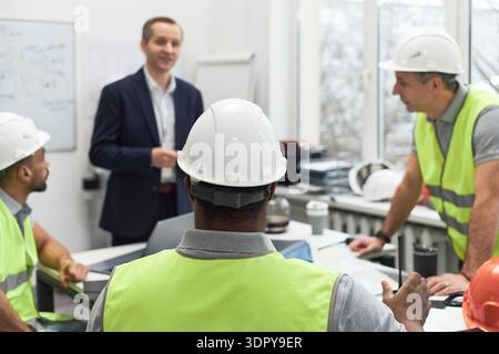 Uomo caucasico di mezza età in piedi e che parla con un gruppo di uomini di mezza età che indossano giubbotti di sicurezza e elmetti in ufficio, team caposquadra che ascolta e discute del progetto Foto Stock