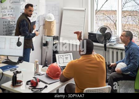 Uomo caucasico di mezza età in piedi vicino alla lavagna, che conduce una discussione con l'uomo nero di mezza età e l'uomo caucasico di mezza età, seduto alla scrivania nella riunione del caposquadra dell'ufficio Foto Stock
