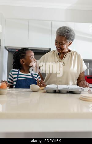 Donna anziana e nipote che cuoce al bancone della cucina di casa con muffin, uova, grembiule in denim Foto Stock