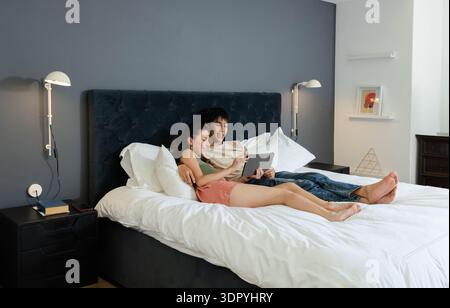Diverse coppie che si reclinano su un grande letto in camera da letto, condividono un tablet vicino alla testata del letto scura Foto Stock