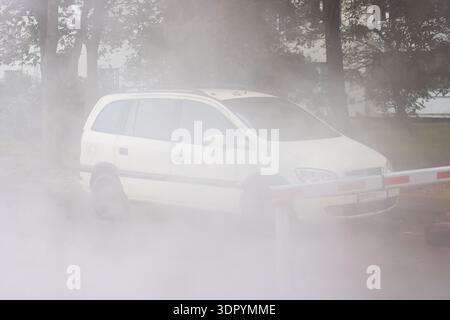 Minivan bianco avvolto dal vapore denso, fermato dietro cancello barriera abbassato, alberi sfocati dalla foschia e pozzanghere riflettenti Foto Stock