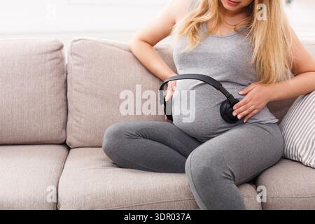 Una donna incinta ascolta la musica con le cuffie sulla pancia mentre è seduta su un divano Foto Stock