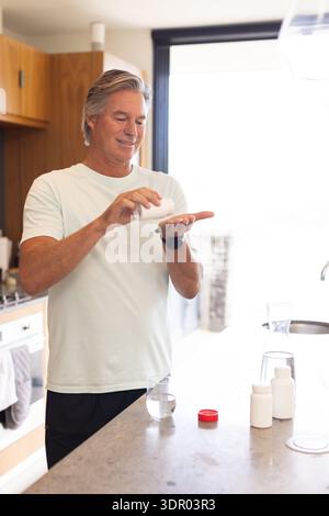 Uomo maturo in piedi a Kitchen Island versando pillole da bottiglia bianca a palma aperta, vetro nelle vicinanze Foto Stock