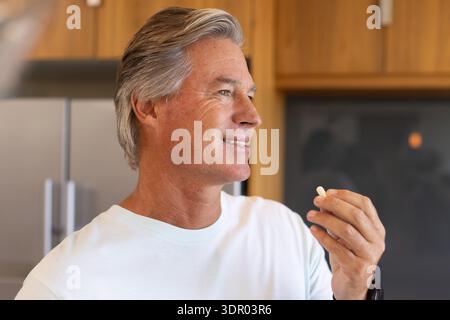 Uomo anziano in piedi in una moderna cucina domestica tenendo una piccola pillola bianca vicino alla bocca con indosso una maglietta bianca Foto Stock