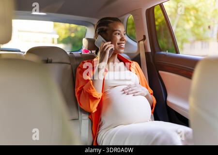 Sorridente giovane donna nera incinta seduta sul sedile posteriore che parla al telefono Foto Stock