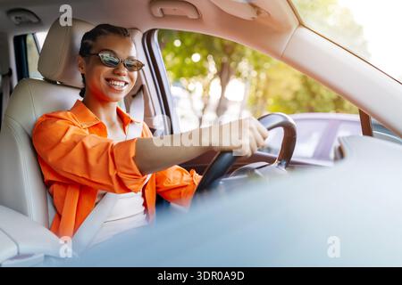 Giovane donna nera che guida un'auto con occhiali da sole, sorridente con un'espressione rilassata Foto Stock