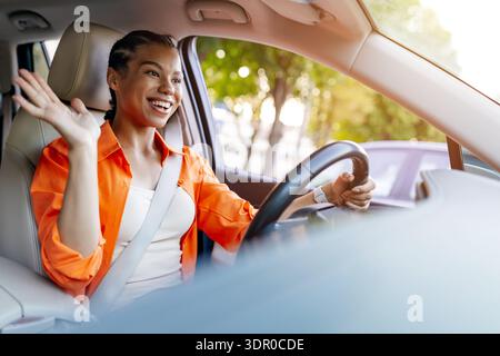 Giovane donna nera che sventola la mano durante la guida dell'auto, esprimendo il saluto Foto Stock