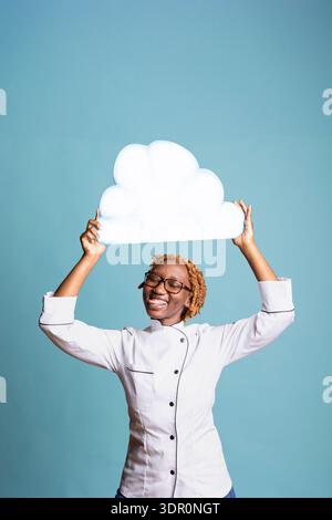 Allegro chef presenta un concetto divertente e creativo per promozioni culinarie, branding di ristoranti o pubblicità di cibo. Una giovane cuoca tiene un cartello nuvola vuoto sopra la testa su sfondo blu. Foto Stock