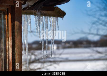 Vista dettagliata di ghiaccioli nitidi e traslucidi formati dalla neve che si scioglie dal vecchio bordo del tetto contro un cielo azzurro e un paesaggio innevato Foto Stock