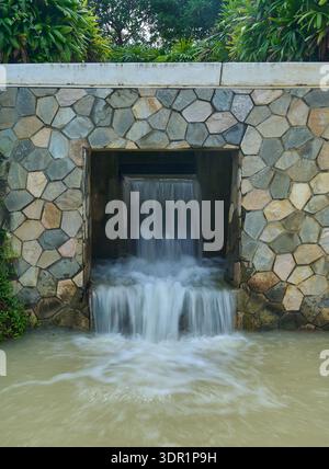 L'acqua che scorre da uno sbocco rettangolare incorporato in un muro di pietra, creando una piccola cascata artificiale. Foto Stock