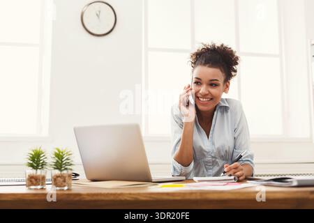 Donna che parla al telefono mentre lavora alla scrivania con notebook in un luminoso spazio ufficio Foto Stock
