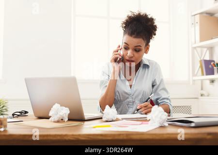 Giovane donna che parla al telefono mentre lavora alla scrivania con notebook e note Foto Stock